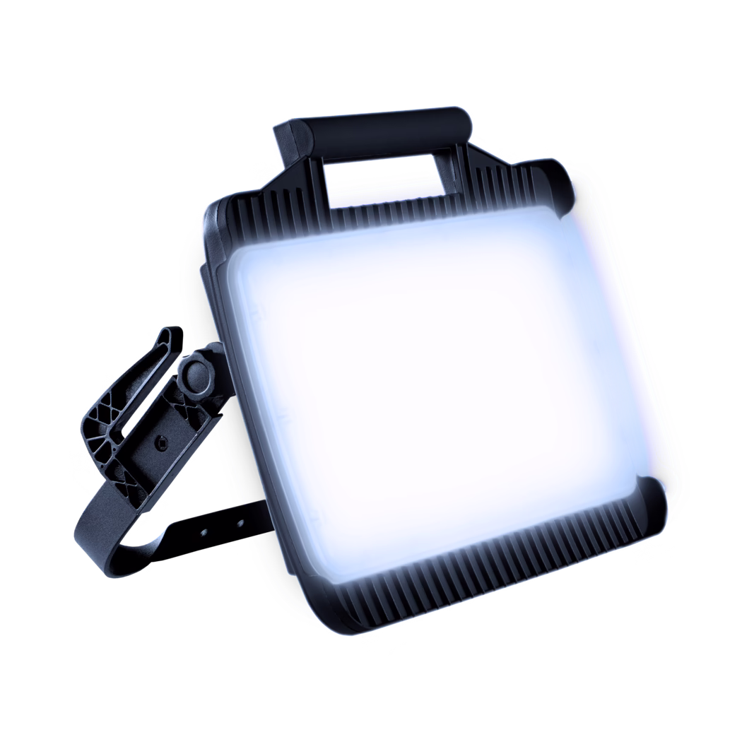 Gros plan d'un projecteur de chantier LED rectangulaire avec une surface lumineuse intense, un boîtier noir à nervures, une poignée supérieure et un support réglable, présenté en angle sur un fond blanc lumineux.