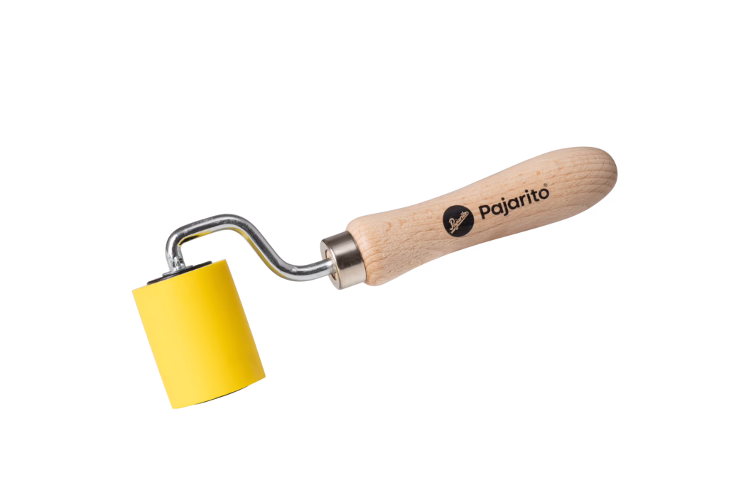 Pajarito_Dummy_Seam controller PU foam Performance Line_034398_2021-02 High-resolution product photograph of a small hand roller tool photographed on a clean white background. The tool features a smooth, light beech-wood handle printed with the brand name "Pajarito" and a round logo in black. A chrome-plated metal shaft with a distinctive bent offset connects the handle to a cylindrical yellow rubber or foam roller. The roller is bright yellow, approximately small-to-medium in diameter, with flat ends and a uniform surface. The composition places the tool diagonally across the frame from lower left toward upper right, showing clear detail: visible wood grain and texture on the handle, subtle metallic reflections on the shaft, and soft shadowing beneath the roller and handle. The lighting is even and neutral, emphasizing the product’s colors and materials without harsh contrasts. This image is suitable for e-commerce, product catalogs, or instructional content and conveys the tool’s size, materials, branding, and intended use for smoothing, pressing or applying even pressure on surfaces such as wallpaper, vinyl, adhesives or printmaking applications.
