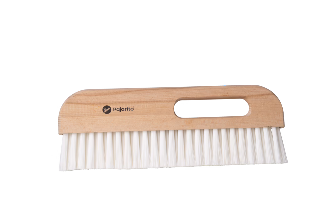 Gros plan d'une brosse à main en bois clair avec une poignée rectangulaire et une ouverture ovale pour le doigt, portant la marque 'Pajarito' imprimée sur le manche ; des poils synthétiques blancs et denses s'étendent droit depuis la base. La brosse est posée à l'horizontale sur un fond blanc neutre et semble propre et inutilisée. Conçue pour dépoussiérer et balayer les petites saletés sur les meubles et les sols, elle associe une prise en bois naturel lisse à des poils résistants pour un nettoyage quotidien.