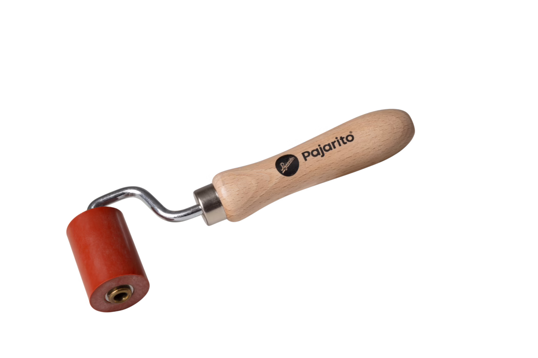 Photo de produit en gros plan d'un petit rouleau à main sur fond blanc. L'outil présente un manche ergonomique en bois clair imprimé du nom de marque « Pajarito », une tige métallique coudée avec une douille métallique et un rouleau cylindrique rouge en caoutchouc avec un moyeu central en laiton. Conception compacte pour lisser ou presser des matériaux.