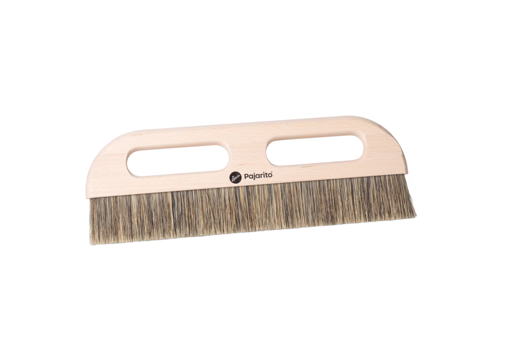 Gros plan d'une brosse à main en bois avec un manche en hêtre clair et des poils naturels denses. Le manche plat et rectangulaire comporte deux ouvertures ovales allongées pour une prise sûre à deux mains et un petit logo noir Pajarito imprimé au centre. Les poils sont constitués de fibres mélangées beige et brune, coupés uniformément en un bord droit, adaptés au balayage de poussières fines et de petits débris sur les sols ou les surfaces de travail. La brosse est photographiée sur un fond blanc propre, mettant en valeur le grain du bois poncé, les coins arrondis et le design ergonomique et pratique.
