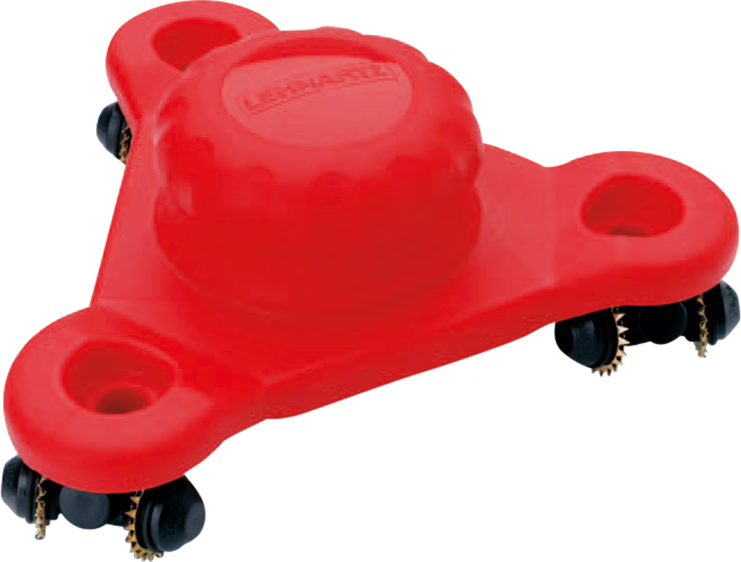 Close-up product shot of a bright red plastic furniture mover skate. The triangular, three-lobed plate features a raised central dome with a molded logo and three recessed circular pockets near each corner. Under the edges are black multi-wheel roller assemblies with small brass-coloured toothed sprockets visible. The image is cropped on a white background and highlights the textured plastic surface, the central dome detail and the roller mechanism used to slide heavy items across floors.