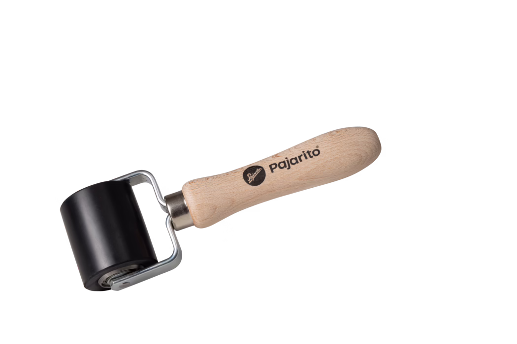 Pajarito_Dummy_Pressure roller small_038700_2021-02 A small hand roller tool photographed on a white background, consisting of a smooth black rubber cylindrical roller mounted on a metal frame and attached to a light beech wood handle stamped with the brand 'pajarito'. The handle is ergonomically shaped, sanded, and shows visible wood grain; the metal bracket connects to a bearing allowing the roller to rotate freely. The tool is isolated, well-lit, and positioned diagonally with the handle on the right and the roller on the left. Suitable for tasks such as applying pressure when laminating, smoothing wallpaper or decals, and printmaking.