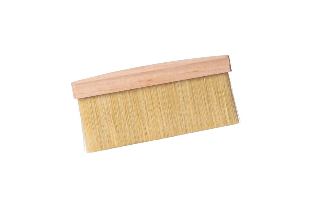 Un large pinceau plat avec une poignée en bois clair et des poils synthétiques beiges et denses, présenté sur fond blanc avec une légère inclinaison. Ce pinceau est généralement utilisé pour la pose de papiers peints, le dépoussiérage ou l'application de vernis ; les poils longs et serrés sont fixés sur une plaque de bois simple et non vernie.